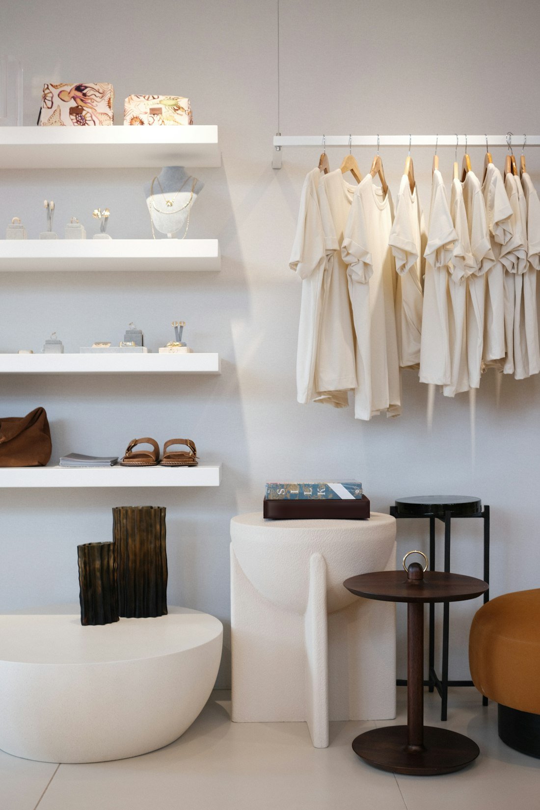 Boutique minimaliste : vêtements crème sur un portant, étagères blanches, tabourets sculptés